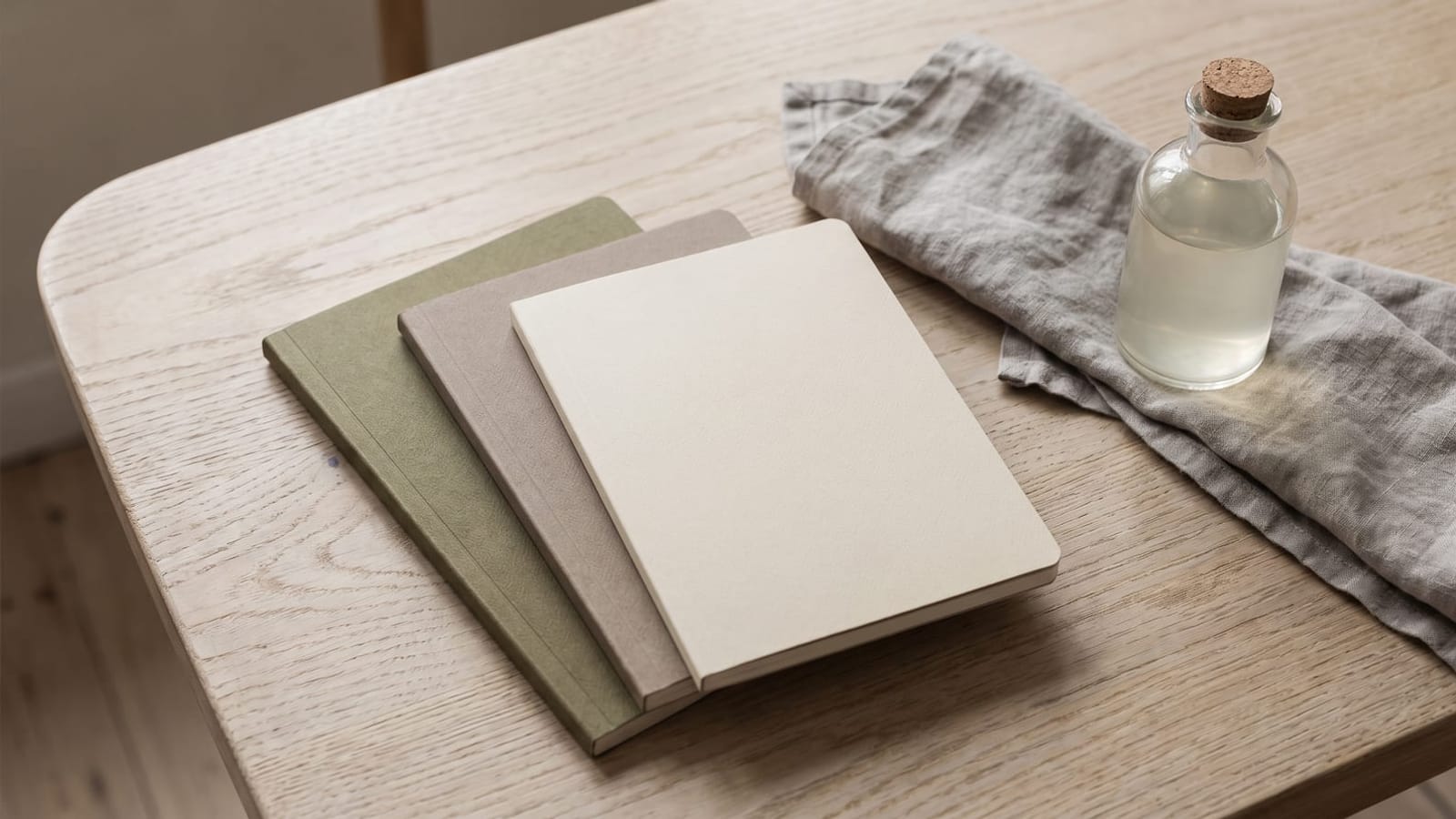 Three closed unlabelled notebooks, a linen napkin and a glass bottle on a light oak table